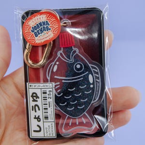 Può includere: Un portachiavi a forma di pesce con glitter liquidi e tappo rosso, riempito di glitter scuri. La confezione include un'etichetta rossa circolare con il testo Joanna Behar Itadakimasu e un'etichetta rettangolare con 25g e caratteri giapponesi.