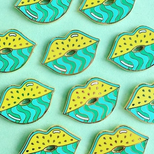Op de afbeelding: Negen emaillepins in de vorm van lippen met een geel en groen ontwerp. De lippen zijn omrand met goud.