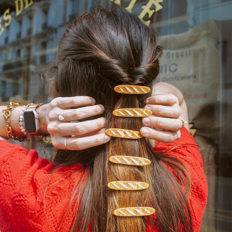 May include: A woman with long brown hair is wearing a red shirt and has multiple gold and white striped barrettes in her hair.