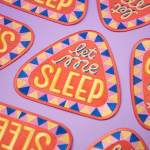 May include: A set of colorful iron-on patches with a triangular shape and a retro design. The patches feature the text "Let me sleep" in a yellow font with pink and blue accents.