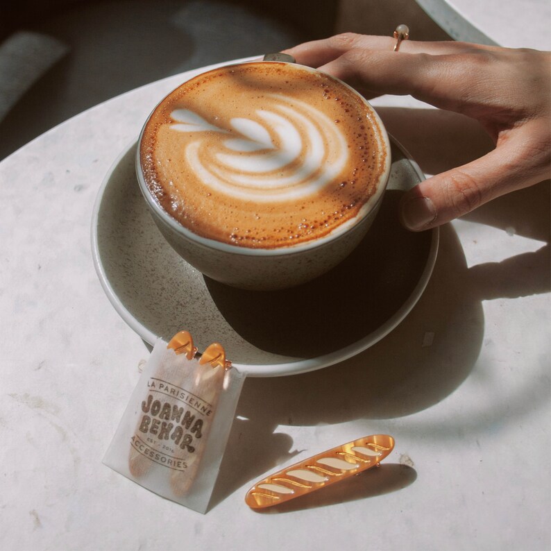 May include: A cup of coffee with latte art in a white ceramic mug with a saucer. The mug is sitting on a white table with a gold hair clip and a small white bag with the text "La Parisienne Joanna Bekar" printed on it.