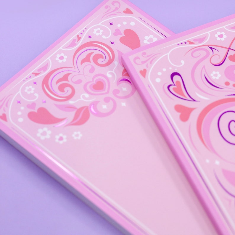 May include: Two pink notebooks with a decorative border of pink, white, and purple swirls and hearts. The notebooks are stacked on top of each other.