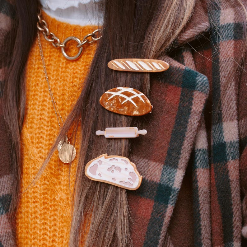May include: A woman with long brown hair wearing a brown and black plaid jacket and a yellow sweater. She has a gold chain necklace and four hair clips in her hair shaped like bread and a rolling pin.