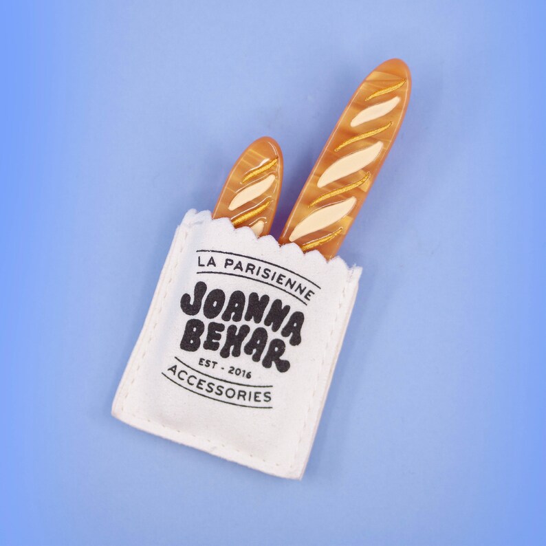 May include: A white fabric bag with the text "La Parisienne Joanna Behar Est. 2016 Accessories" printed on it. Two brown baguette-shaped hair clips are sticking out of the top of the bag.