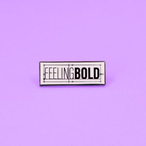 May include: White enamel pin with black outline and text that reads "FEELING BOLD".