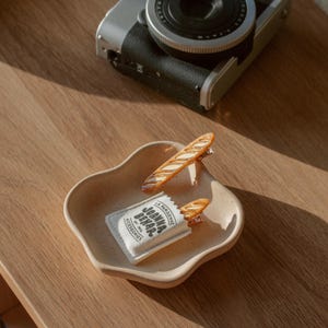 May include: A white fabric bag with the text "La Parisienne Joanna Behar Est. 2016 Accessories" printed on it. Two brown baguette-shaped hair clips are sticking out of the top of the bag in a wavy plate on a wooden surface