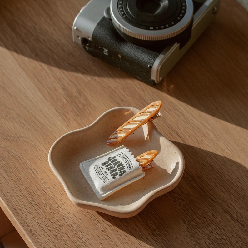 May include: A white fabric bag with the text "La Parisienne Joanna Behar Est. 2016 Accessories" printed on it. Two brown baguette-shaped hair clips are sticking out of the top of the bag in a wavy plate on a wooden surface
