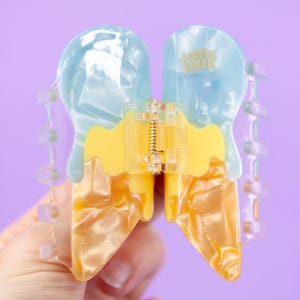 May include: A fun, ice cream cone-shaped hair clip. The clip features a light blue and yellow design with a clear, decorative edge. The clip has the text "joanna behar" in gold lettering.