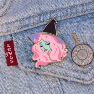 K&ouml;nnte beinhalten: Eine goldfarbene Emaille-Anstecknadel mit einer Cartoon-Hexe mit rosa Haaren, einem lila Hut und einem gr&uuml;nen Gesicht. Die Hexe hat die Augen geschlossen und einen ruhigen Ausdruck. Die Anstecknadel ist vor einem Jeans-Hintergrund platziert.