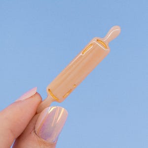 Puede incluir: Un pequeño rodillo de amasar de color rosa claro con detalles dorados. El rodillo se sostiene en una mano con esmalte de uñas rosa claro.