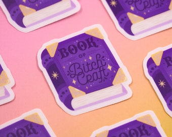 Puede incluir: Pegatina de libro morado y dorado con el texto "Book of Bitch Craft" en la portada. La pegatina tiene un borde blanco y está sobre un fondo rosa.
