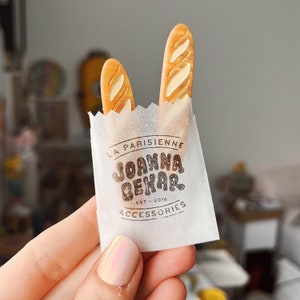 May include: A white paper bag with the text "La Parisienne Joanna Behar Est. 2016 Accessories" printed on it. Two brown baguette-shaped hair clips are sticking out of the top of the bag.