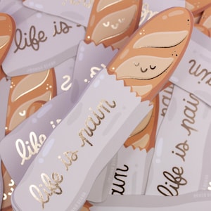 Può includere: Un set di carte con sfondo viola chiaro e lettere dorate. Le carte presentano un'illustrazione di baguette e il testo "life is pain".