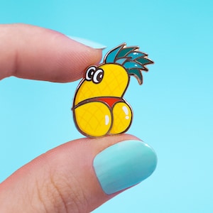 May include: A yellow pineapple enamel pin with a red bikini bottom. The pineapple has a face with black eyes and a green and blue leafy top. The pin is held between two fingers against a light blue background.