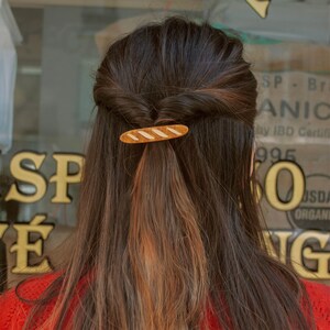 May include: A woman with long brown hair with a light brown hair clip shaped like a baguette. The hair clip is in the middle of her hair.