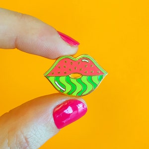 Op de afbeelding: Een gouden emaille pin in de vorm van lippen met een watermeloenontwerp. De bovenlip is rood met zwarte pitten en de onderlip is groen met witte strepen.