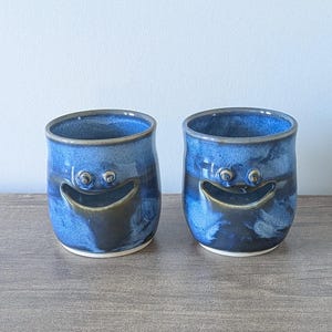 May include: Two blue ceramic cups with a smiling face design. The cups have a dark blue glaze with lighter blue accents and a dark brown mouth. The cups are on a wooden surface.