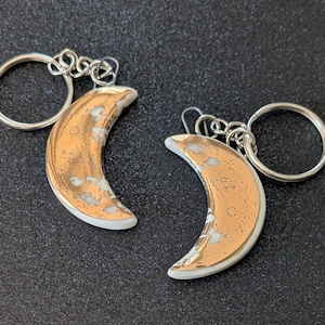 May include: Two crescent moon keyrings. The moons have a gold metallic finish with white accents and are attached to silver keyrings. The keyrings are displayed on a dark grey surface.