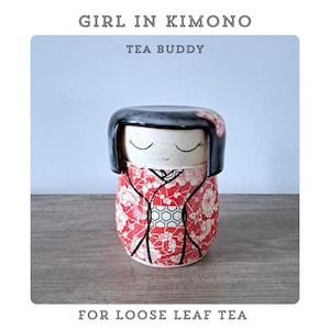 May include: A ceramic tea buddy shaped like a girl in a kimono. The tea buddy features black hair and a red and white floral kimono design. The text "GIRL IN KIMONO", "TEA BUDDY", and "FOR LOOSE LEAF TEA" are displayed.