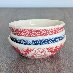 May include: Three small ceramic bowls stacked on top of each other. The top bowl has a red floral pattern. The middle bowl has a blue floral pattern. The bottom bowl has a red floral pattern.