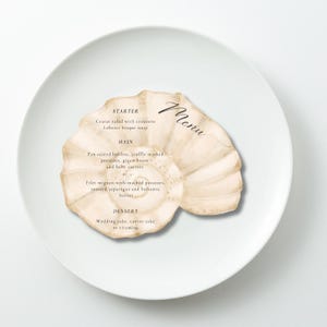 May include: A seashell-shaped menu card with handwritten "Menu" script. The menu lists starter, main, and dessert options, including Caesar salad, filet mignon, and wedding cake. The card is on a white plate.