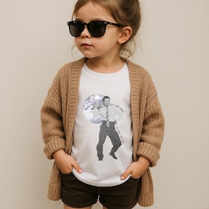 Può includere: Un bambino indossa una maglietta bianca con una grafica in bianco e nero di un uomo in giacca e cravatta, una palla da discoteca e la scritta "Dudes Don't Dance, No More They Say". Il bambino indossa anche occhiali da sole neri, un cardigan beige e pantaloncini marroni.