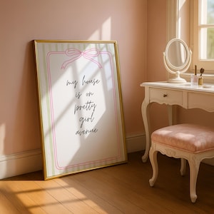 Puede incluir: Un cuadro enmarcado con el texto "my house is on pretty girl avenue". El cuadro tiene un marco dorado, un lazo rosa y un borde a rayas rosa y verde. El cuadro está apoyado contra una pared rosa en una habitación con una cómoda.