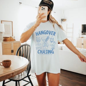 Puede incluir: Camiseta azul claro con las palabras "HANGOVER CHASING" en azul, junto con ilustraciones de cócteles, gafas de sol y otros elementos de fiesta. El texto "Carolina's Bachelorette Weekend 2024" también está impreso en la camiseta.