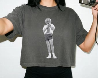 May include: A charcoal gray cropped t-shirt featuring a black and white graphic print of a person wearing a helmet, a collared shirt, a tie, and shorts. The person is standing and holding an object near their face. The shirt has short sleeves.