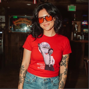 以下が含まれることがあります： 明るい赤の半袖Tシャツ。ライトピンクのドレスシャツとネクタイを着用し、頭上にミラーボールを掲げた男性のグラフィックプリントが施されている。グラフィックの下には、白い大文字で「RESPECT YOUR MOTHER」というテキストがプリントされている。
