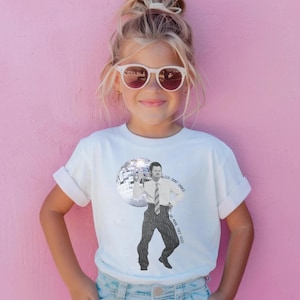 May include: A white short-sleeved t-shirt featuring a black and white graphic of a person in a dress shirt and pinstripe trousers standing next to a disco ball. The text "DJS DONT DANCE NO MORE THEY SAY" is printed in a curved arc next to the figure.