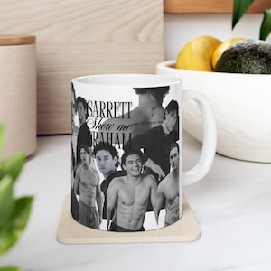 Può includere: Tazza in ceramica bianca con un collage di immagini in bianco e nero di una persona. La tazza ha il testo "GARRETT Show me GRAHAM" stampato sopra. La tazza è su un sottobicchiere beige.