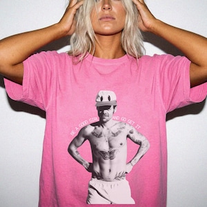Puede incluir: Una camiseta de manga corta de color rosa brillante con un estampado gráfico en blanco y negro de una persona sin camisa que lleva una gorra de béisbol. El texto "BE A GOOD GIRL AND GO GET IT" aparece impreso en arco sobre el gráfico.