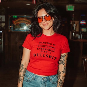 Könnte beinhalten: Ein leuchtend rotes Kurzarm-T-Shirt mit dem schwarzen Aufdruck DANCING IS EVERYTHING EVERYTHING ELSE IS BULLSHIT. Das T-Shirt wird mit einer blauen Jeans kombiniert und mit orange getönten Sonnenbrillen mit schwarzem Rahmen getragen.