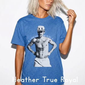 Pode incluir: Camiseta azul Heather True Royal com uma imagem em preto e branco de uma pessoa usando boné e shorts, com tatuagens. O texto "BE A GOOD GIRL AND GO GET IT" está impresso acima da imagem. As palavras "Heather True Royal" estão impressas na parte inferior.