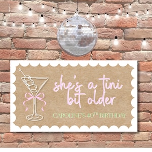 May include: A rectangular sign with a scalloped edge, featuring a martini glass illustration and the text "she's a tini bit older". The sign also includes the text "CAROLINE'S 40TH BIRTHDAY". A disco ball and string lights hang above the sign against a brick wall.