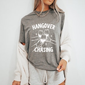 May include: A gray t-shirt with the words "HANGOVER CHASING" in white, along with an illustration of three wine glasses clinking. The shirt is short-sleeved and made of a soft material.