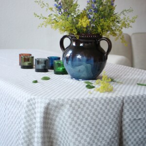 Puede incluir: Un jarrón de cerámica azul y marrón con un ramo de flores silvestres moradas y amarillas sobre un mantel a cuadros blancos.