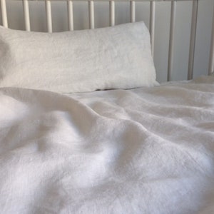 Puede incluir: Una sábana de cuna de lino blanco y una almohada, con la sábana metida alrededor de la almohada. La sábana está arrugada y tiene un aspecto natural y texturizado.