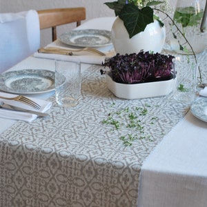 Peut inclure: Une nappe blanche avec un chemin de table beige à motifs. Un vase blanc avec des feuilles vertes et des fleurs blanches est placé au centre de la table. Deux verres en verre transparent et deux assiettes blanches avec des bordures dorées sont sur la table. Un petit bol blanc avec des pousses violettes est placé devant le vase.