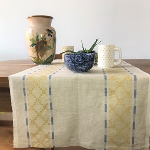 Könnte beinhalten: Ein hellbeiger Tischläufer aus Leinen mit gewebten gelben und blauen Streifen. Der Läufer liegt auf einem Holztisch mit einer blauen und weißen Keramikschüssel, einer weißen Keramiktasse mit weißen Punkten und einer braunen Keramikvase mit Blumendesigns.
