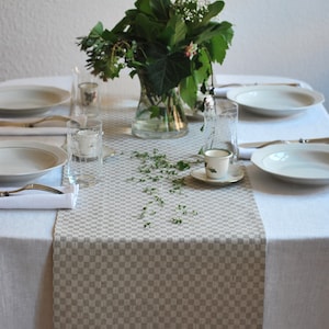 Op de afbeelding: Een beige geblokte tafelkleed met een boeket groene bladeren en witte bloemen in een glazen vaas op een witte tafelkleed. Twee witte borden en twee glazen staan op de tafel.
