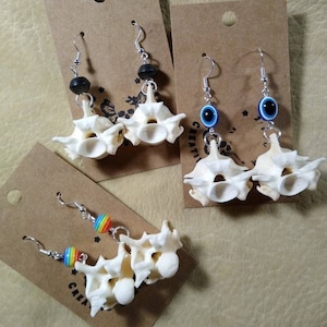 May include: Three pairs of earrings featuring bone vertebrae with different colored beads. The first pair has black beads, the second pair has blue evil eye beads, and the third pair has rainbow beads.
