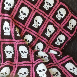 Puede incluir: Una manta de ganchillo con un patr&oacute;n de calavera en blanco y rosa sobre un fondo marr&oacute;n.