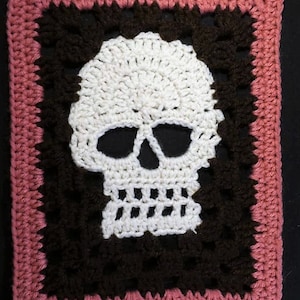 Puede incluir: Una calavera de ganchillo con la cara blanca y los ojos negros, enmarcada por un borde de ganchillo marr&oacute;n y un borde exterior de ganchillo rosa.