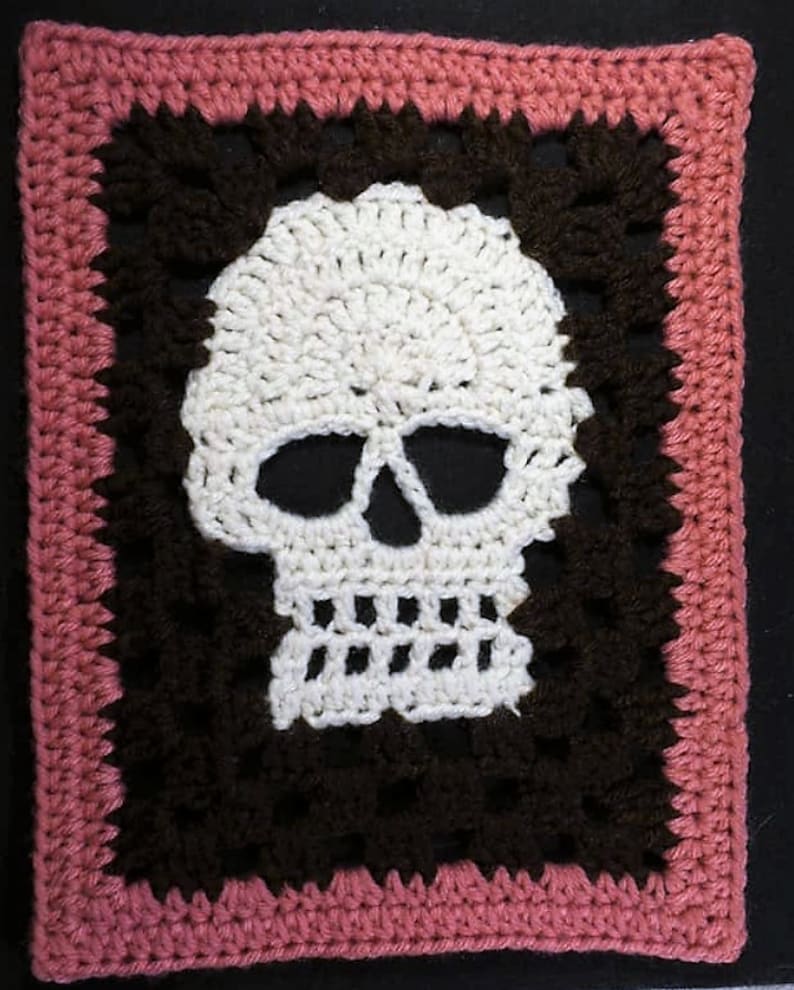 Puede incluir: Una calavera de ganchillo con la cara blanca y los ojos negros, enmarcada por un borde de ganchillo marr&oacute;n y un borde exterior de ganchillo rosa.