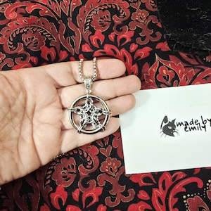 May include: Silver-toned pendant necklace featuring a pentagram design within a circle, with a chain detail. The necklace is displayed on a red and black patterned fabric. A small card with a cat logo and the text "made by emily" is also visible.