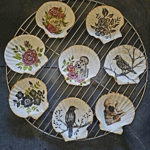 May include: Seven decorative seashells with painted designs. The designs include a skull with roses, a crow on a branch, a skull with a skeleton, black flowers, a skull, and two different floral designs.