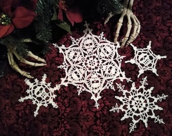 May include: Four white crocheted snowflakes with skull designs on a dark red background with a skeletal hand.