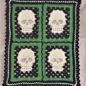 Puede incluir: Una manta de ganchillo con un esquema de color negro, verde y blanco. La manta presenta cuatro dise&ntilde;os de calaveras.